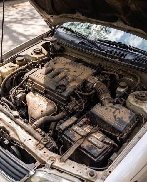 An open car engine bay dusty and grimy with road film across the covers and hoses.