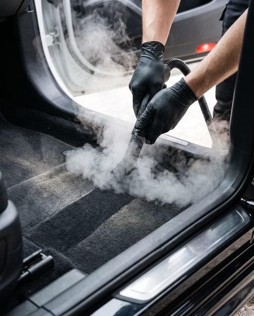 A detailer running a steam wand across a car carpet with visible vapour.