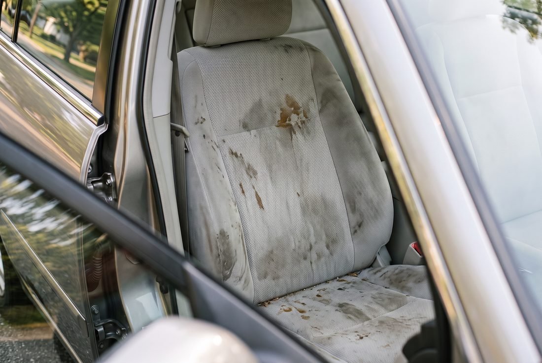 A light grey cloth car seat heavily soiled with several dark set-in stains and a greyed surface.